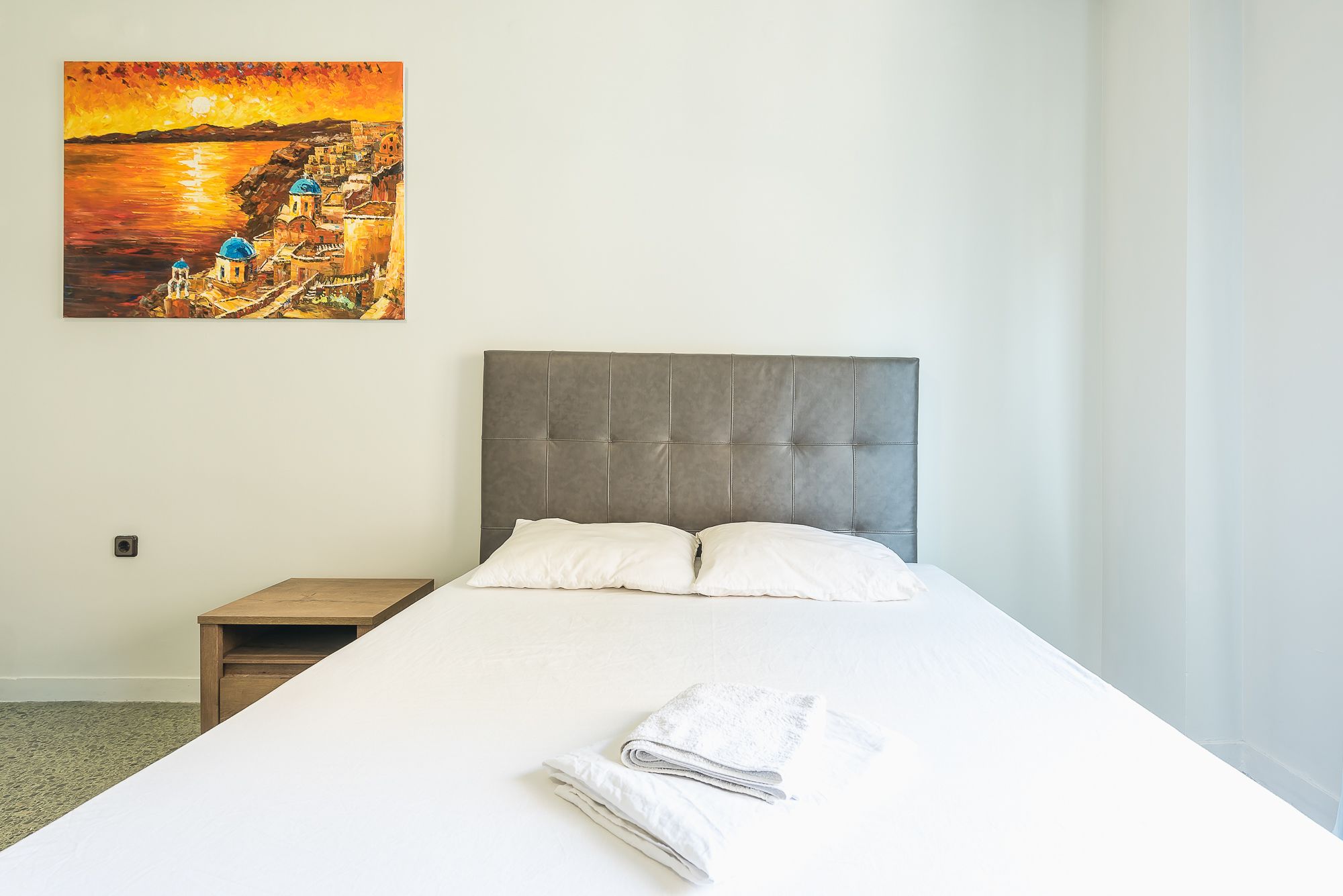 A hotel room with a large bed, a grey upholstered headboard, and a painting of a coastal scene on the wall.