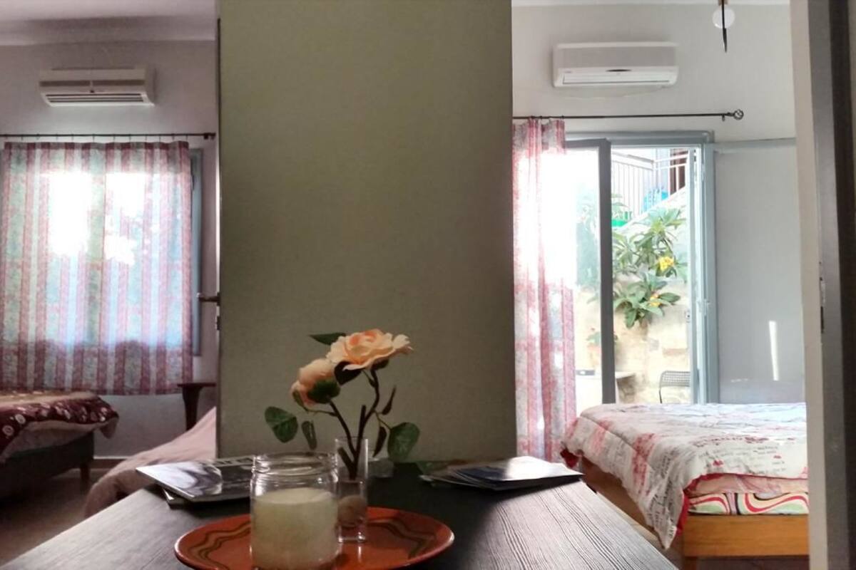 A room with a bed, a small table with a candle, a vase with a single flower, and a window with curtains.