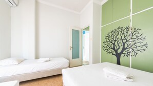 Une chambre d’hôtel avec deux lits, un mur vert orné d’une fresque représentant un arbre, et une porte donnant sur une autre pièce.