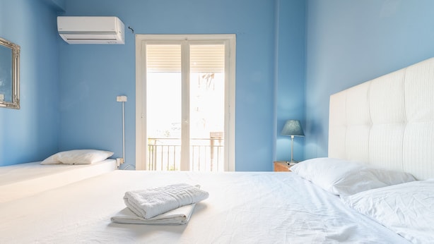 Une chambre avec un grand lit, un plus petit lit, une fenêtre avec des stores, une table de chevet avec une lampe et un appareil de climatisation.