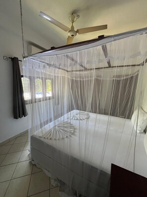 Una habitación con una cama cubierta por una mosquitera, un ventilador de techo y una ventana con cortinas.