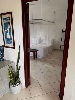 Una habitación con una cama cubierta con sábanas blancas, una mesita pequeña, una planta en maceta y una escalera apoyada contra la pared.