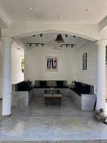 Un espacio de estar al aire libre cubierto, con una mesa central, bancos acolchados y un ventilador de techo.