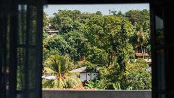 Vista a través de una ventana con balcón, con un paisaje verde exuberante y follaje denso, y algunos edificios al fondo.
