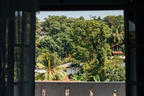 Une vue à travers une fenêtre donnant sur un balcon, avec un paysage verdoyant et luxuriant, une végétation dense et quelques bâtiments en arrière-plan.