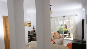 Ein Wohnzimmer mit einer Couch, einem Couchtisch und einer TV-Konsole. Eine Tür führt in einen anderen Raum, ein Fenster mit Vorhängen ist ebenfalls zu sehen.