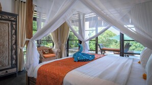 A bedroom with a bed, a wooden wardrobe, a bench, and a view of greenery through large windows.