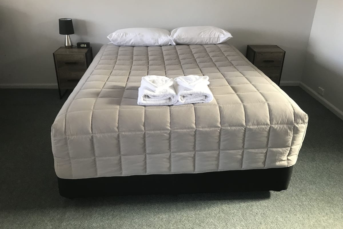 A neatly made bed with a beige quilted bedspread and two white pillows. There are two bedside tables with lamps and a small clock on one. The room has a carpeted floor and plain walls.
