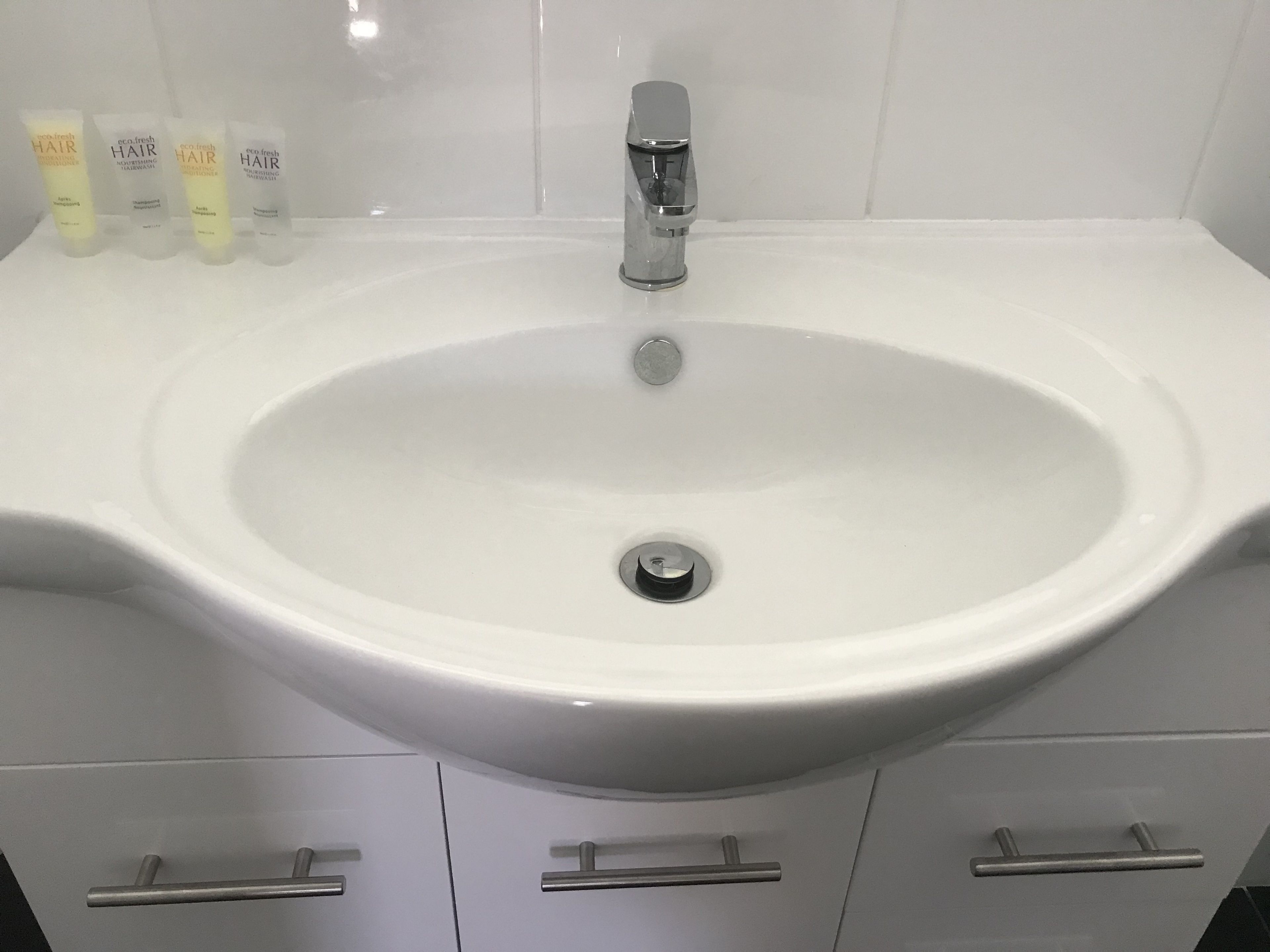 Een witte wastafel met een chromen kraan, vier flessen haarverzorging en drie lades eronder.