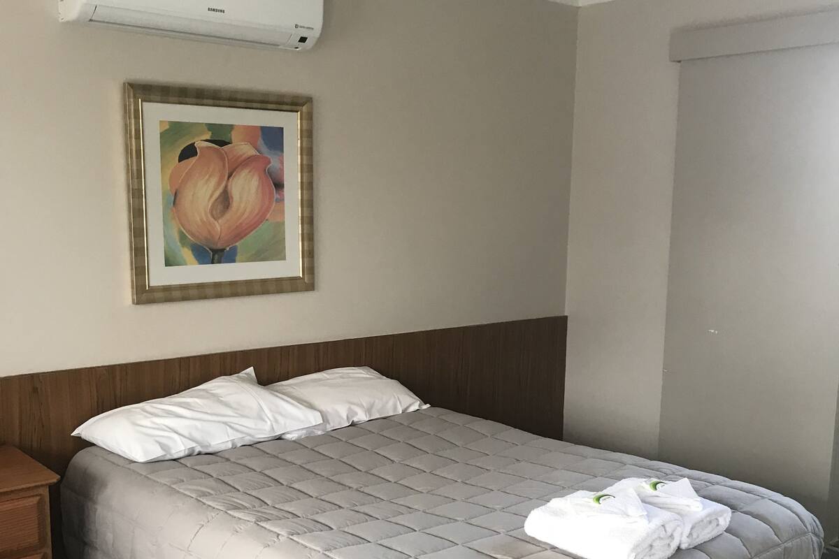 A hotel room with a bed, a nightstand, a painting, and an air conditioning unit.