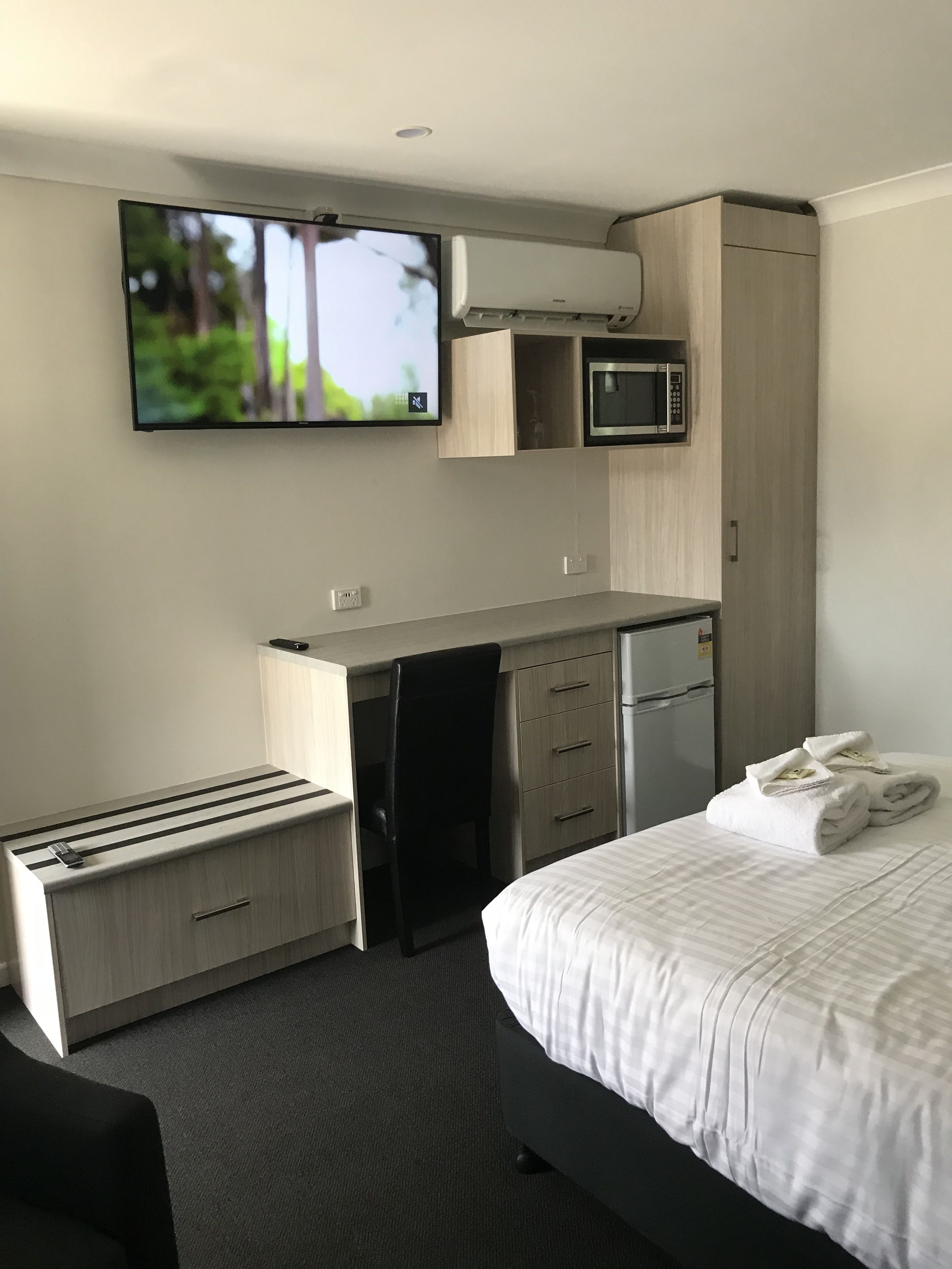 Una habitación de hotel moderna con cama, escritorio con microondas, televisor y una pequeña kitchenette.
