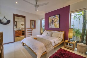 Un dormitorio con cama, ventilador de techo, ventana grande con plantas, mesita de noche de madera y alfombra roja.
