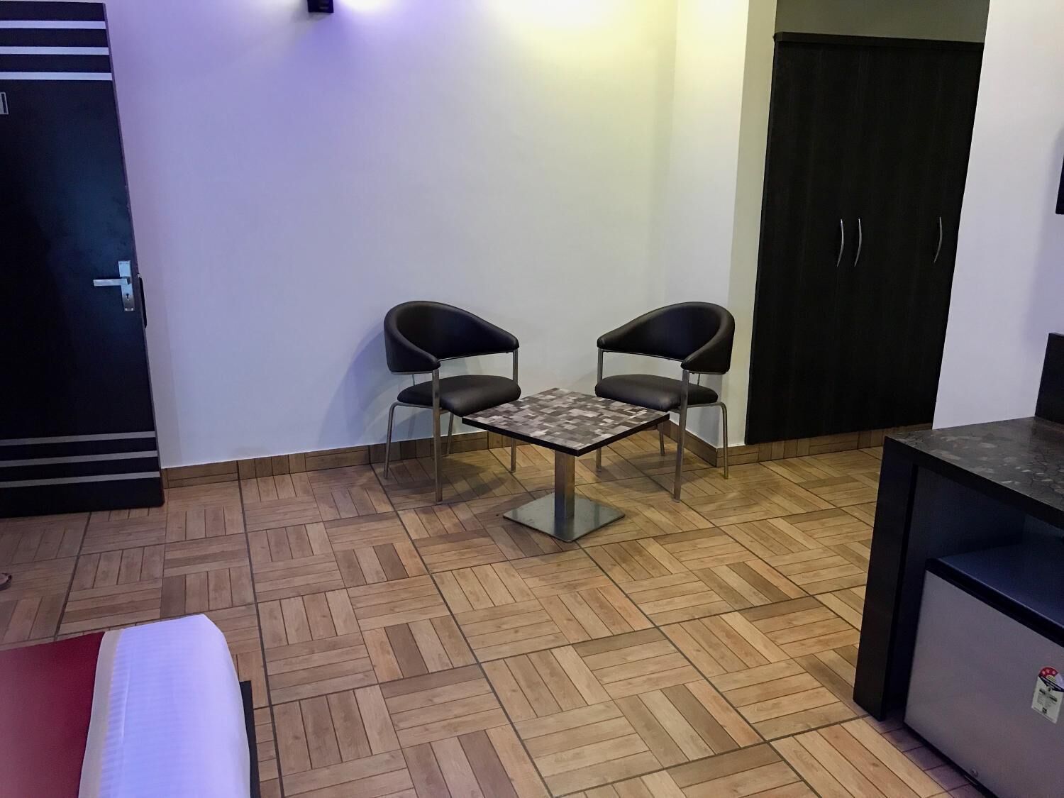 A room with a herringbone-patterned wooden floor, a small table with two black chairs, a dark-colored wardrobe, and a red sofa.