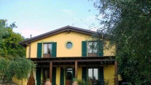 A two-story building with a yellow facade and green shutters, featuring a covered porch and outdoor seating area.