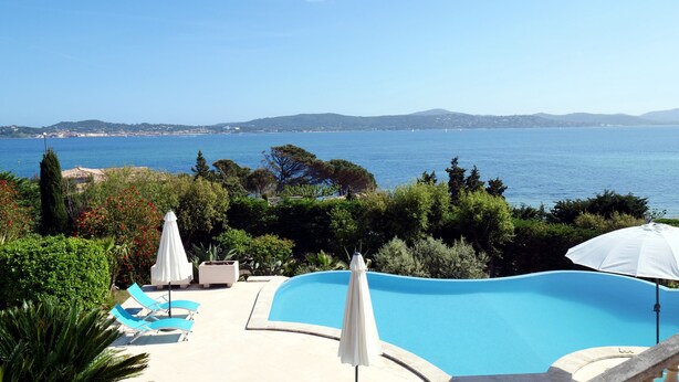 Una piscina con vista al mar, rodeada de vegetación y sillones de descanso.