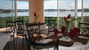 A dining area with tables set for two, large windows offering a view of a lake and greenery. - Ryan's Bay Hotel (Mwanza)