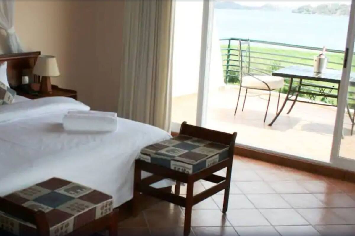 Ein Hotelzimmer mit Bett, Nachttisch mit Lampe, Sessel mit gemustertem Kissen und Balkon mit Tisch und Stuhl.