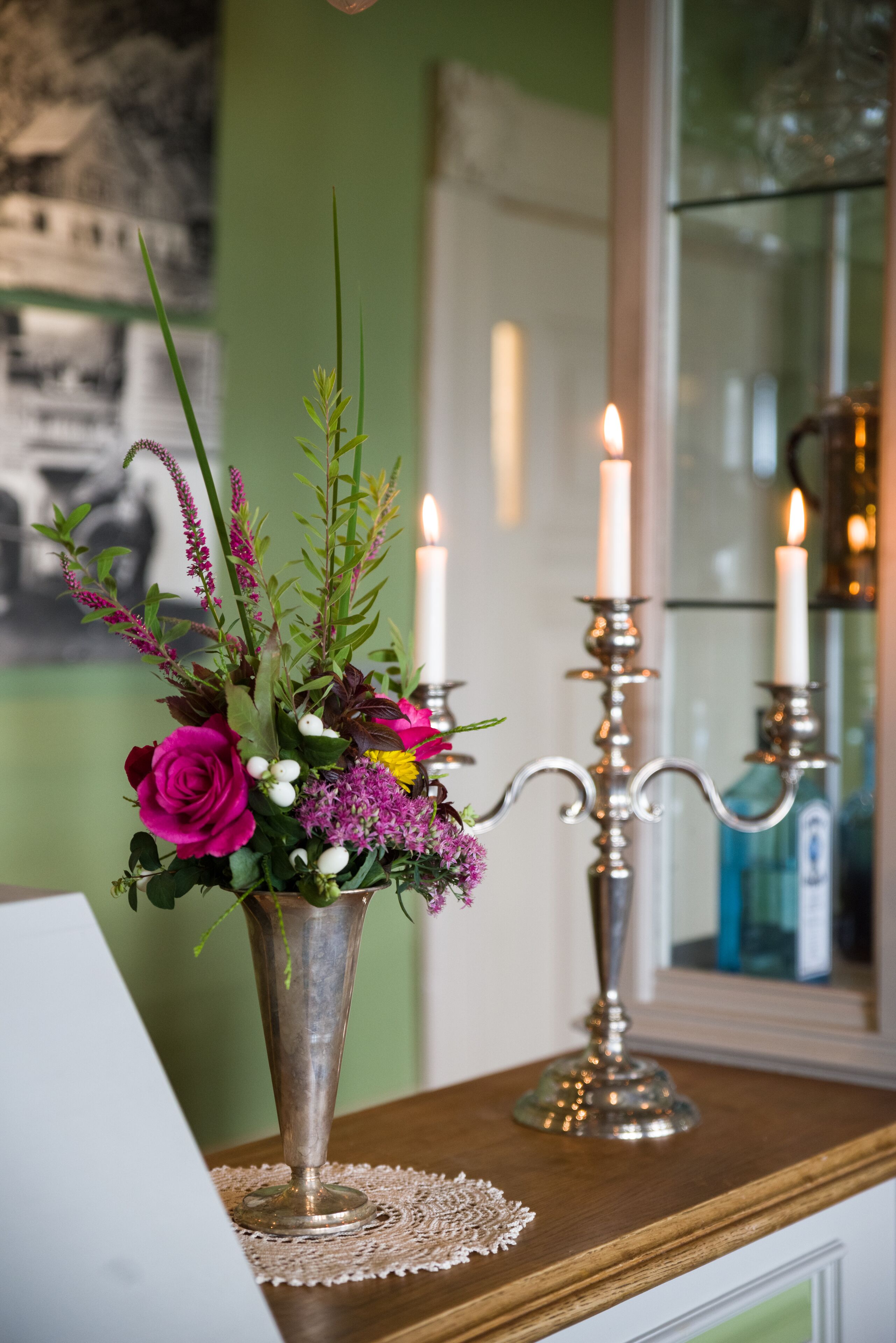 A table with a metal vase containing flowers and a candle holder with two lit candles.