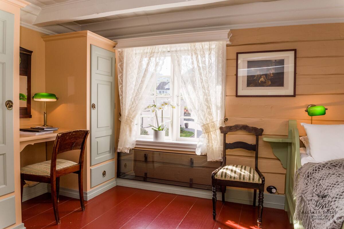 A room with a desk, two chairs, a bed, a window with lace curtains, a framed picture on the wall, and a green lamp.