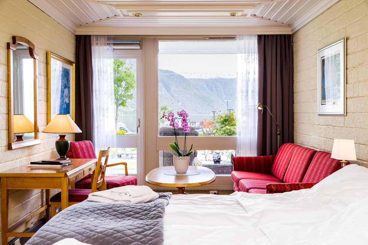 A hotel room with a bed, a red sofa, a small table with a potted plant, a desk with a lamp, and a window with a mountain view.