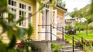 A yellow building with a staircase leading to a door, surrounded by greenery and a small pond. - Walaker Hotel (Luster)