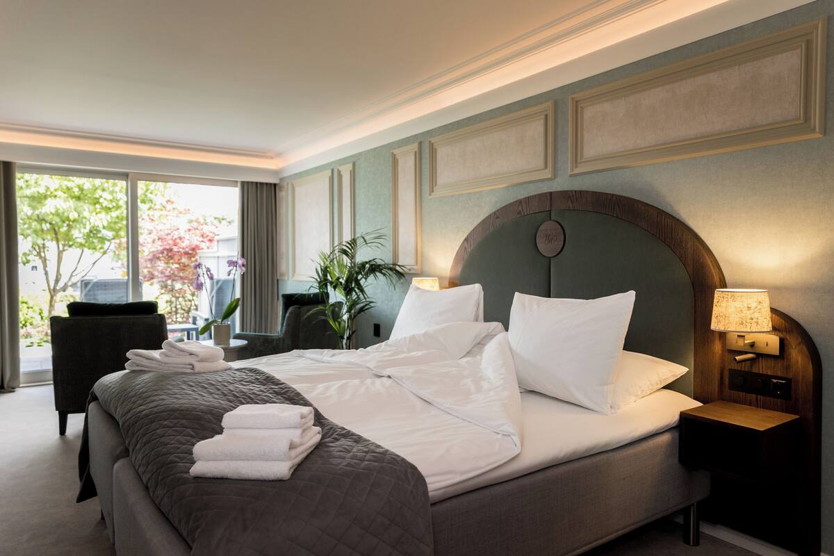 A neatly made bed with white linens, a dark quilted bedspread, and a stack of folded towels. There is a wooden nightstand with a lamp, a dark armchair, and a potted plant beside the bed.