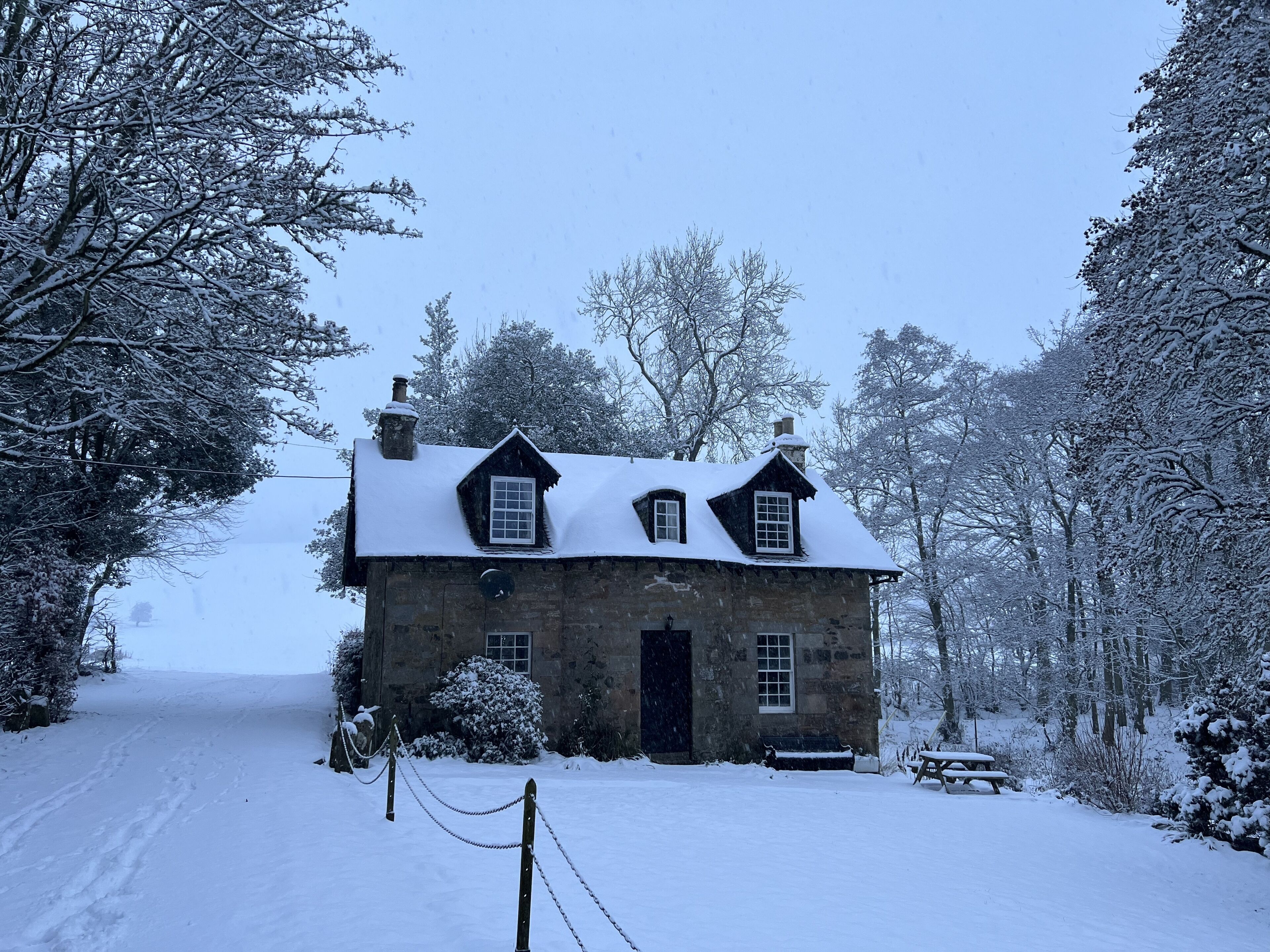 A stone house with a snow-covered roof, surrounded by snow-laden trees.