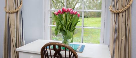 A white desk with a vase of pink tulips, a wooden chair, and a window with a view of a green landscape.