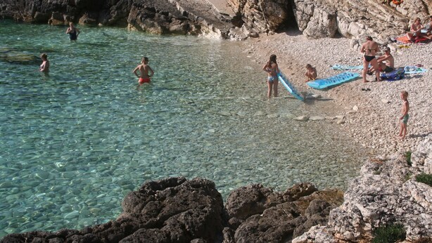 A rocky beach with clear water, people swimming and sunbathing, and a waterfall in the background. - Villa Bramasole ,big pool , View 200m sea, Kamenjak Park, EV charging available (Premantura)