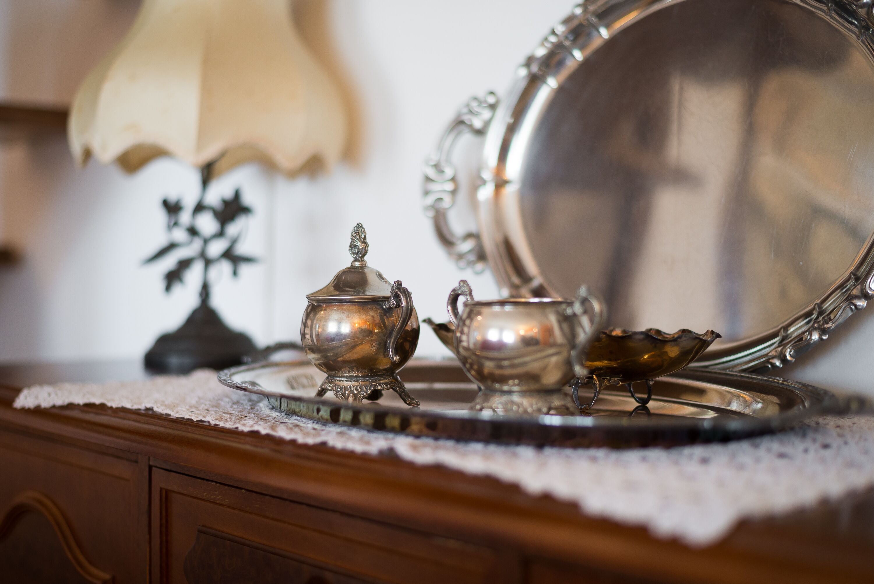 A silver tea set on a lace doily, a lamp, and a mirror on a wooden dresser.