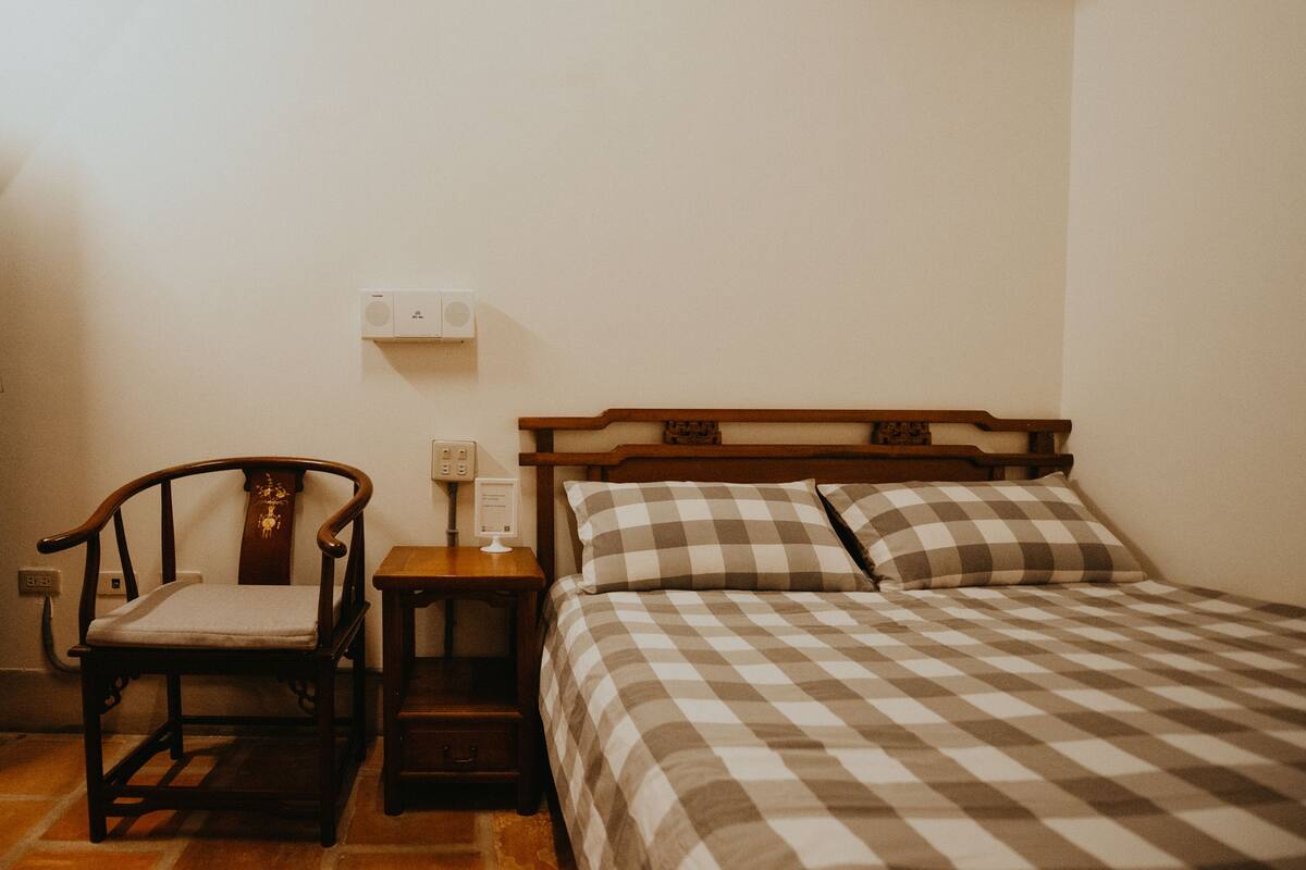 A bedroom with a wooden bed, a checkered bedspread, a wooden chair, and a nightstand with a lamp.