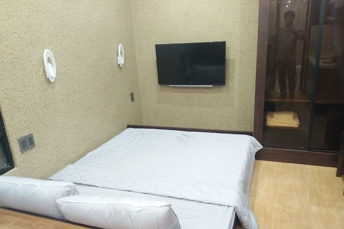 A room with a bed, a television mounted on the wall, and a glass door leading to a bathroom.