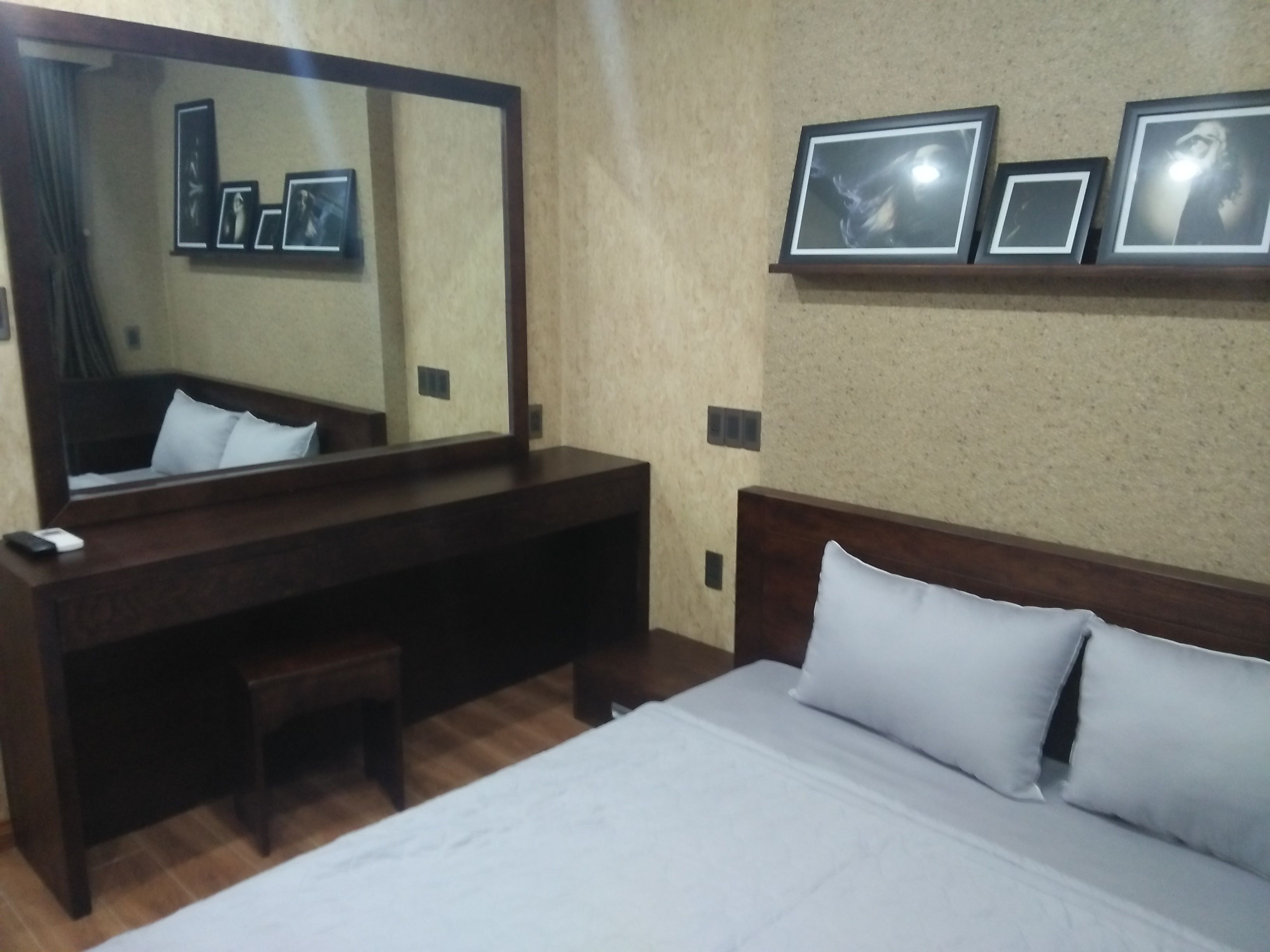 A hotel room with a bed, a large mirror, a wooden desk, and a wall-mounted shelf with framed pictures.