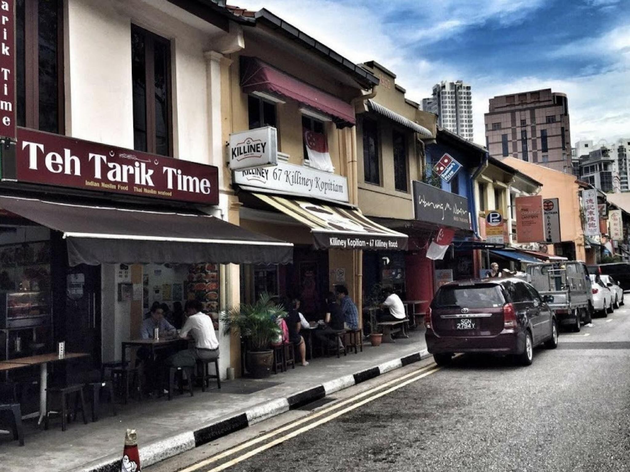 Una calle con diversas tiendas, incluyendo un restaurante llamado 'Teh Tarik Time' y 'Killiney Kopitiam'.