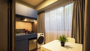Cocina compacta equipada con microondas, tostadora y lavadora. Una pequeña mesa con una planta se encuentra frente a una ventana con cortinas.