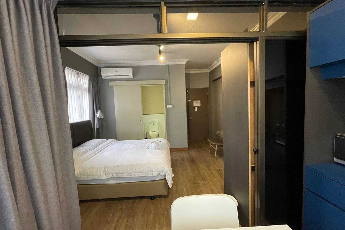 Une chambre à coucher de taille modeste, comprenant un lit, un bureau et une chaise. On peut apercevoir la salle de bain à travers une porte ouverte.