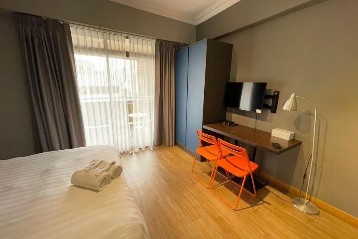 Quarto de hotel com uma cama, uma escrivaninha com duas cadeiras laranja, uma televisão e um abajur.