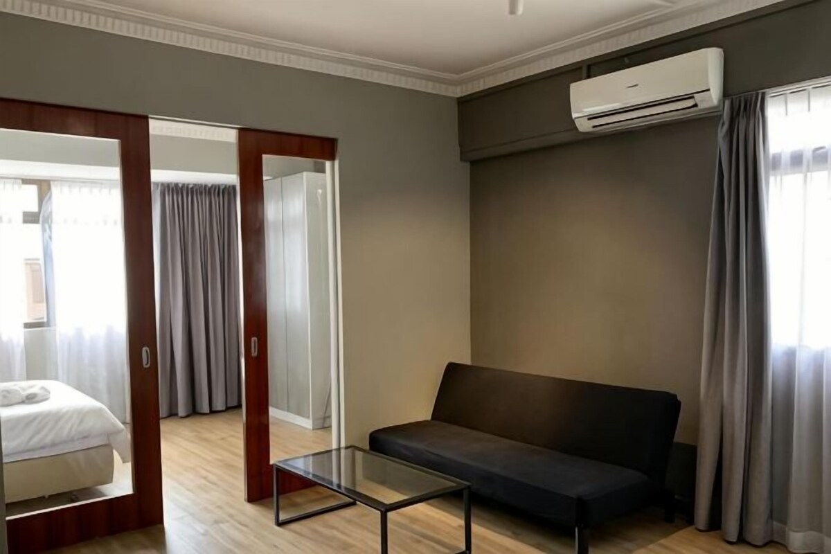 Quarto com sofá, mesa de vidro, cama e porta deslizante que dá para outro cômodo.