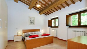 Un dormitorio con cama de madera, mesitas de noche, radiador, ventana con vistas y techo con vigas a la vista.