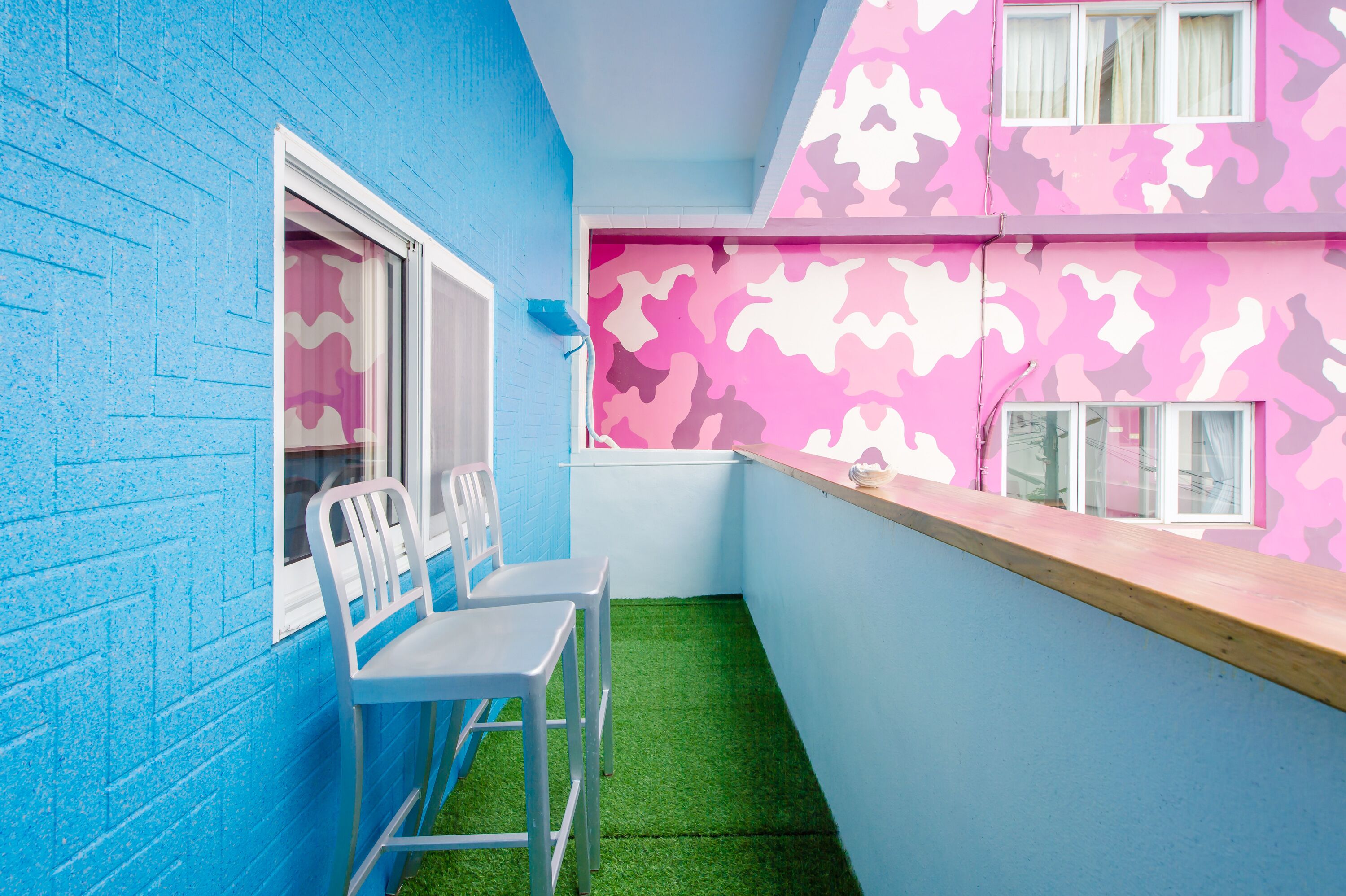 A small, brightly colored room with a blue wall, a pink camouflage-patterned wall, and a green artificial grass floor. There are two white chairs and a small table.