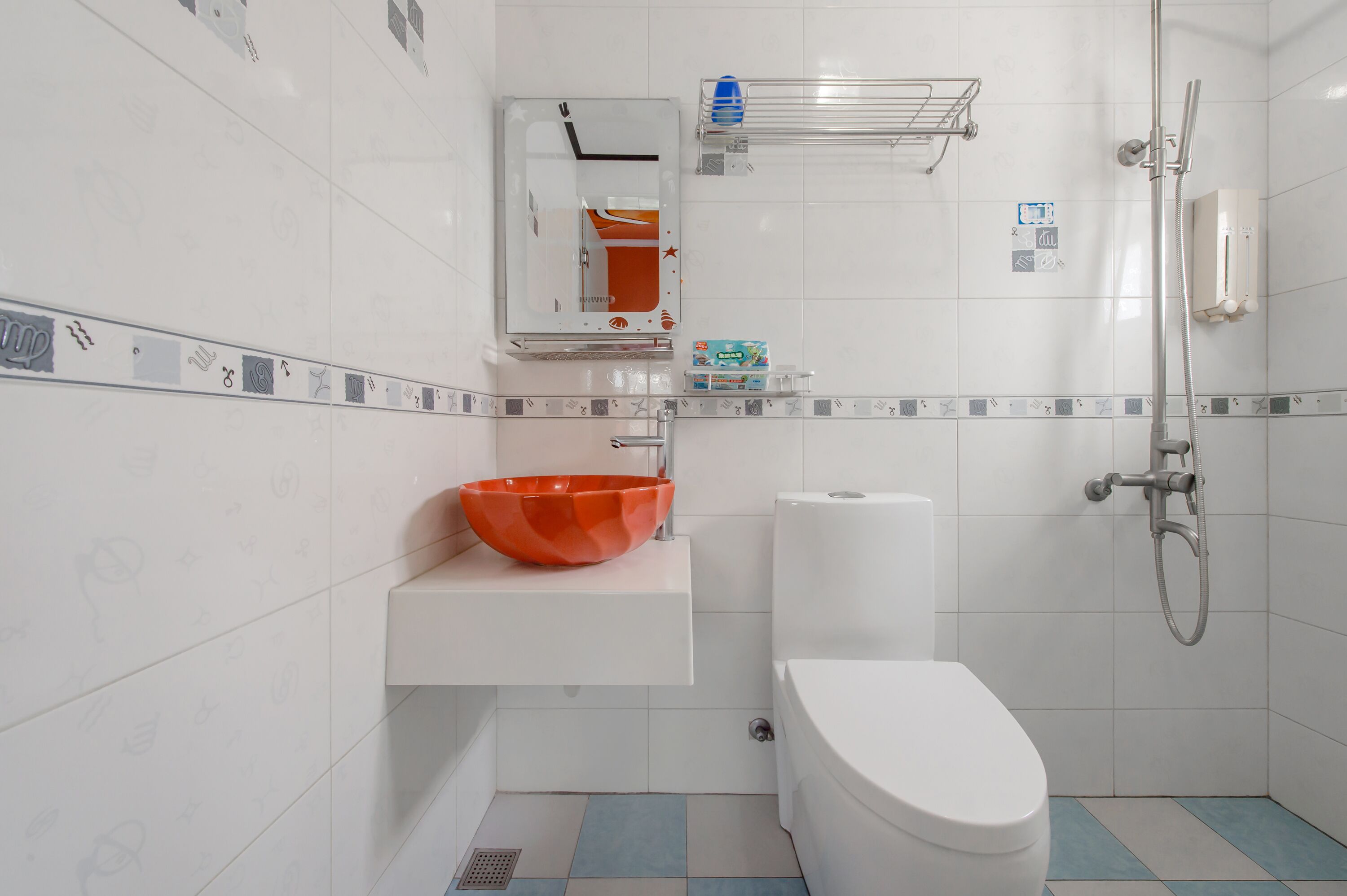 Et badeværelse med et hvidt toilet, en hvid vask med en orange skål, en bruser og et spejl over vasken.