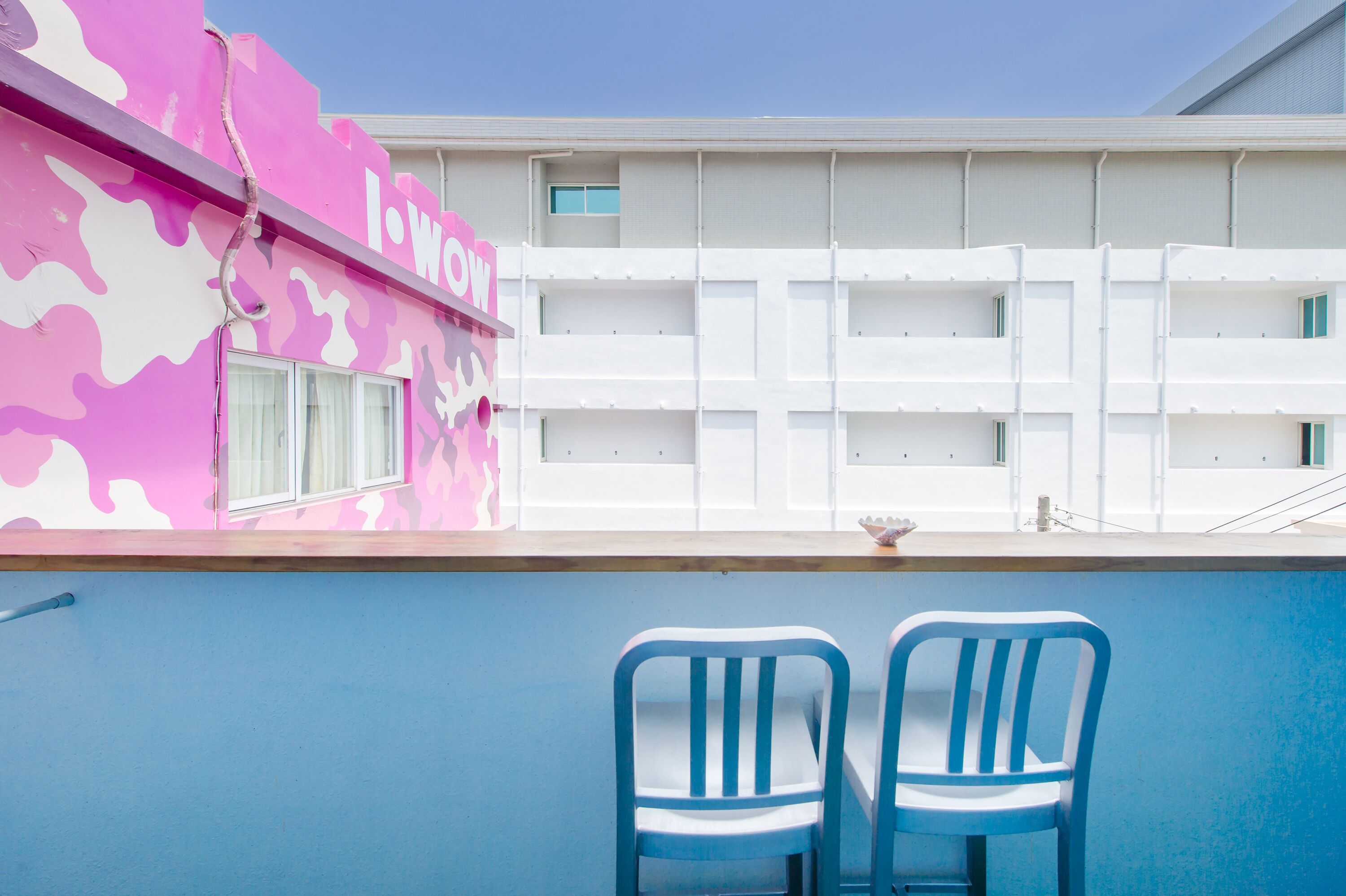 A bar area with two blue chairs, a pink wall with 'low' written on it, and a white building with multiple windows in the background.