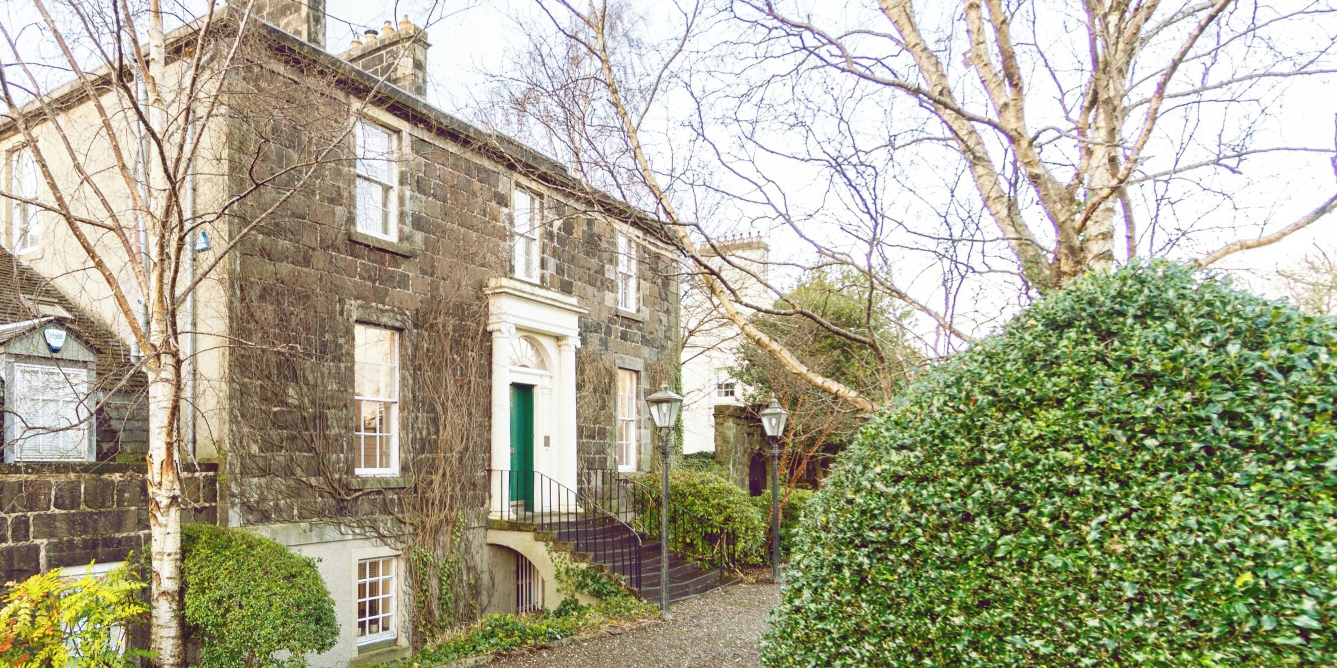 A stone building with a green door, a staircase, and a hedge in the foreground.