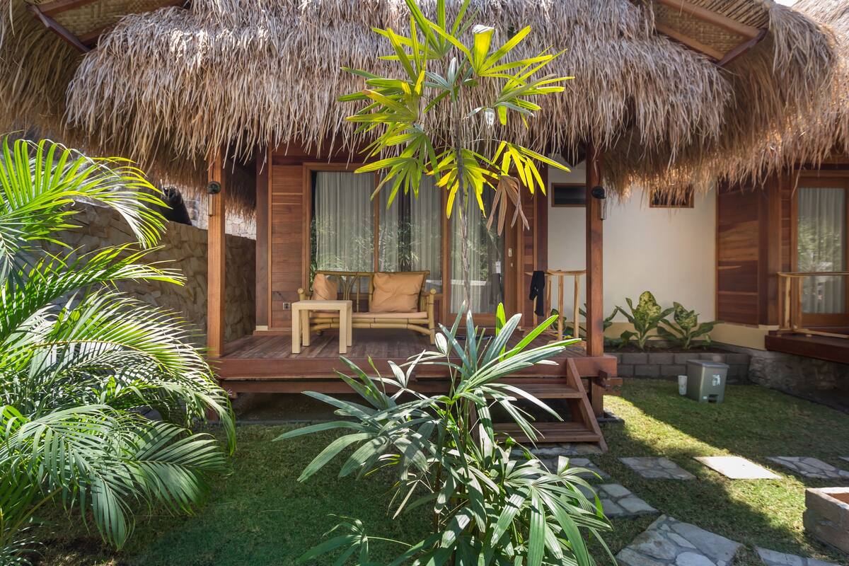 A thatched-roof bungalow with a wooden deck, a sofa, and a small table.