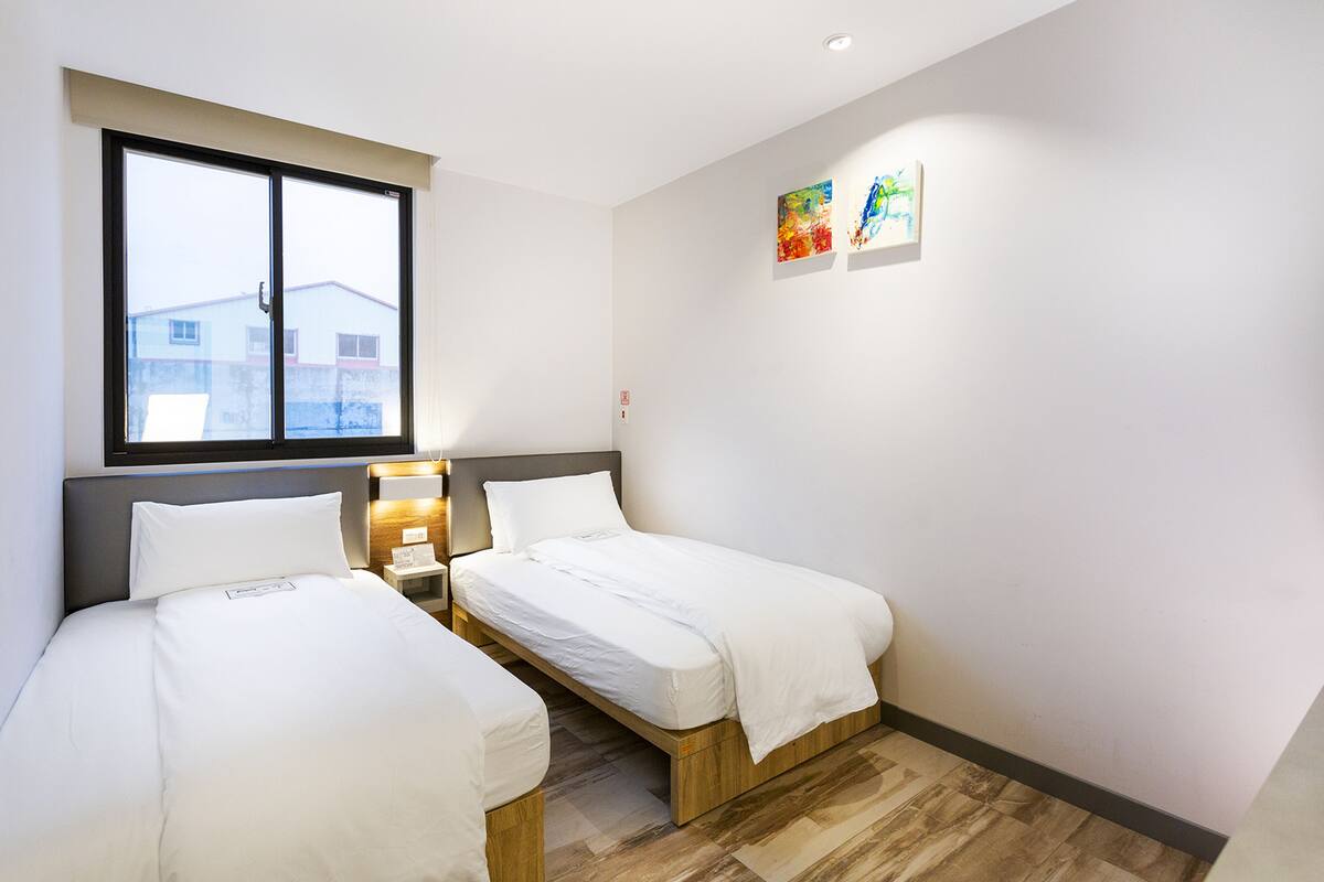 A hotel room with two single beds, a large window, a bedside table with a lamp, and two wall-mounted artworks.