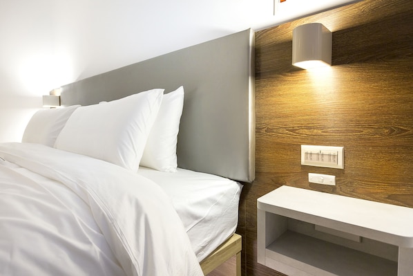 A neatly made bed with white linens, a wooden headboard, a bedside table with a lamp, and a wall-mounted thermostat.
