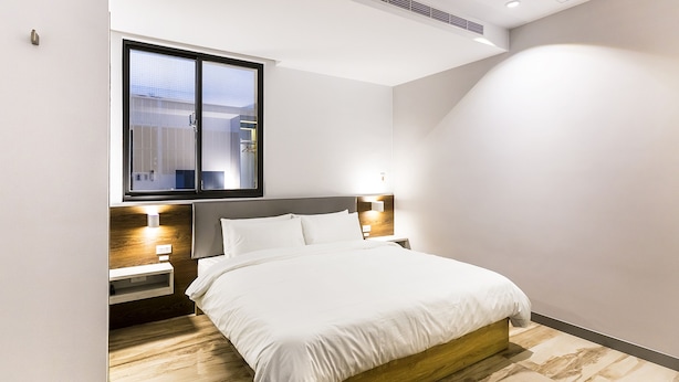 Une chambre d’hôtel moderne dotée d’un grand lit, d’une tête de lit en bois, de tables de chevet avec des lampes et d’une grande fenêtre laissant entrer la lumière naturelle.