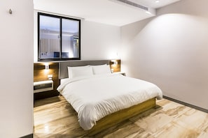 A modern hotel room with a large bed, wooden headboard, bedside tables with lamps, and a large window providing natural light.