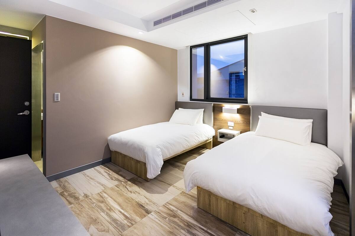 A hotel room with two single beds, a nightstand with a lamp, a large window, and a door to the left.