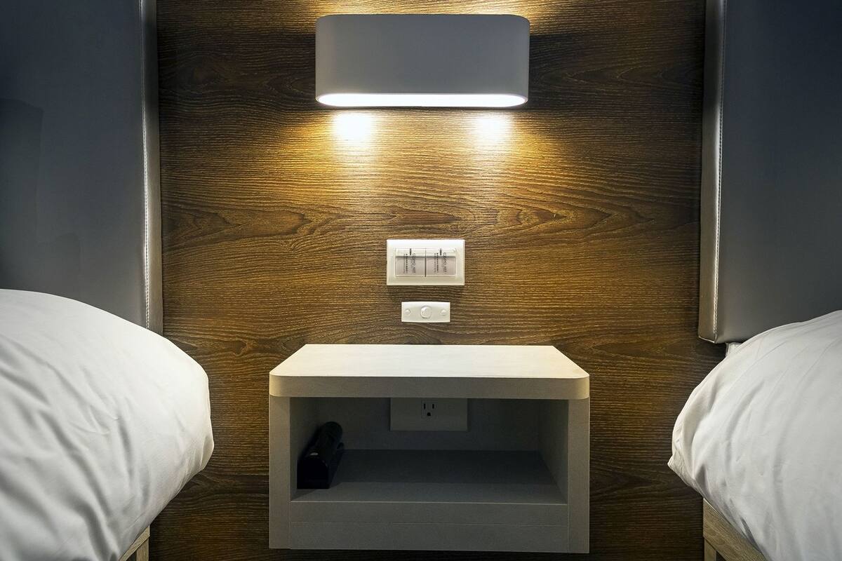 A hotel room with a wooden wall, two beds, a nightstand with a lamp, and a thermostat.